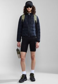 Marine pufferjack, zwarte shorts, groene rugzak, zonnebril, zwarte laarzen met gele veters en een zilveren waterfles.