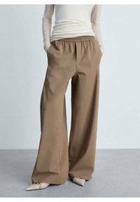 Pantalon marron à jambes larges avec une ceinture élastique, en tissu lisse. Il possède des poches latérales et s'arrête juste au-dessus des chaussures à bout pointu.