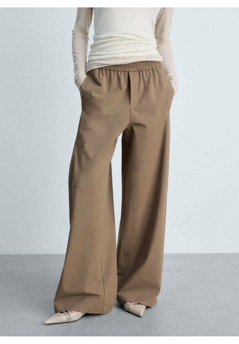 Pantalon marron à jambes larges avec une ceinture élastique, en tissu lisse. Il possède des poches latérales et s'arrête juste au-dessus des chaussures à bout pointu.