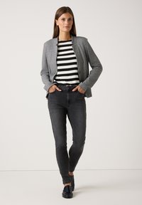 Frau trägt ein schwarz-weiß gestreiftes Hemd, einen grauen karierten Blazer, dunkelgraue Skinny-Jeans und schwarze Slipper, steht mit den Händen in den Taschen.