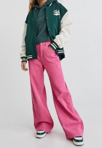 Chaqueta universitaria verde y blanca, sudadera con capucha gris debajo, pantalones anchos de pana rosa y zapatillas con tono verde. Conjunto audaz y casual.