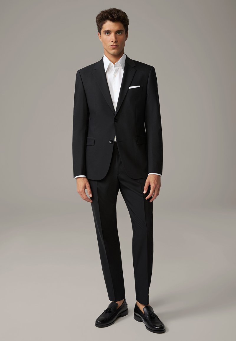 Completo nero con rever a punta, abbinato a una camicia bianca e scarpe nere. Presenta linee pulite e un design slim fit.