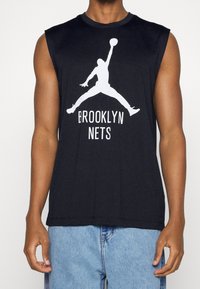 Zwarte mouwloze shirt met wit logo van een springende basketbalspeler en de tekst "Brooklyn Nets", gedragen door iemand in blauwe spijkerbroek.