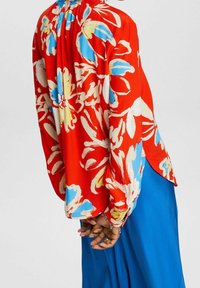 Blouse rouge à motifs floraux bleus, jaunes et blancs. Manches longues avec poignets froncés ; dos avec plis et ourlet arrondi.