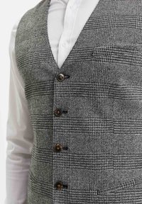 Grå mönstrad väst med en strukturerad vävdesign, med V-hals, fem bruna knappar och en sidoficka. Bärs över en vit skjorta.