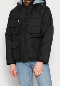 Musta pehmustettu takki, jossa vetoketjullinen suljenta, neljä etutaskua, säädettävä huppu ja raidallinen vuori. Sileä pinta ja kevyt muotoilu.