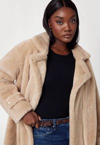 Bež plašček iz umetnega krzna z širokim ovratnikom in gumbastimi manšetami, prelit čez črno majico, v kombinaciji z temnimi kavbojkami in vzorčastim pasom.