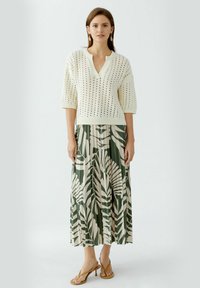 Un pull au crochet blanc avec un décolleté en V et des manches courtes, associé à une longue jupe plissée verte ornée d'un motif de feuilles blanches. Sandales marron.