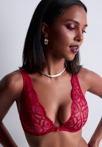Soutien-gorge en dentelle rouge avec un motif géométrique, décolleté plongeant, bords festonnés et un accent doré au centre. Bretelles ajustables.