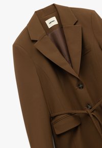 Brun blazer lavet af glat stof, der har en klapkrave, to forreste knapper og et taljebånd. Indeholder een brystlomme og en sidelomme.