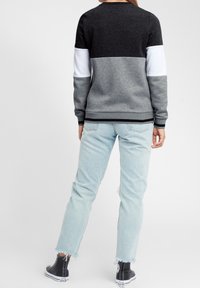 Mujer vestida con un suéter a rayas en negro, blanco y gris, jeans claros con dobladillo deshilachado y zapatillas altas negras, de pie con la espalda hacia la cámara.