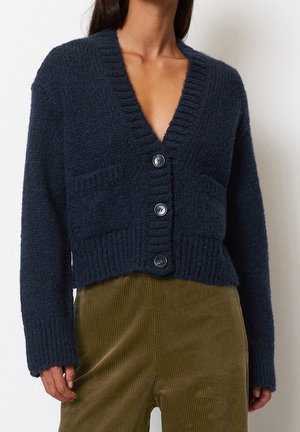 Frau trägt eine dunkelblaue, gestrickte, geknöpfte Strickjacke mit Vorderentaschen und olivgrünen Cordhosen, gezeigt von der Brust bis zur Hüfte.