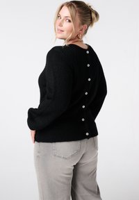 Zwart gebreide trui met lange mouwen, een ronde halslijn en een knoopdetail aan de achterkant. Gecombineerd met grijze spijkerbroek.