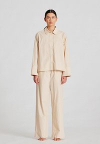Set din bumbac bej, format dintr-o  cămașă cu guler și pantaloni largi, având închidere cu nasturi și mâneci lungi cu o croială lejeră.