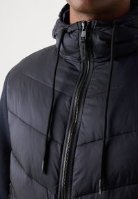 Navy puffer vest with a high collar, zippered front, and adjustable drawstrings. Features stitched panels and a smooth, glossy texture.