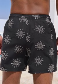 Costume da bagno nero con motivi di soli bianchi, vita elasticizzata e una tasca posteriore con chiusura a scatto. Il tessuto appare liscio e leggero.