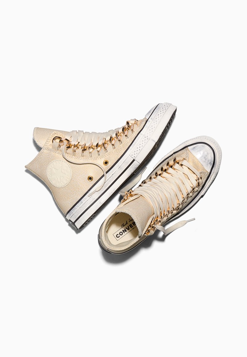 High-top beige Converse sneakers with gold chain eyelets and white satin laces placed side by side on a white background.