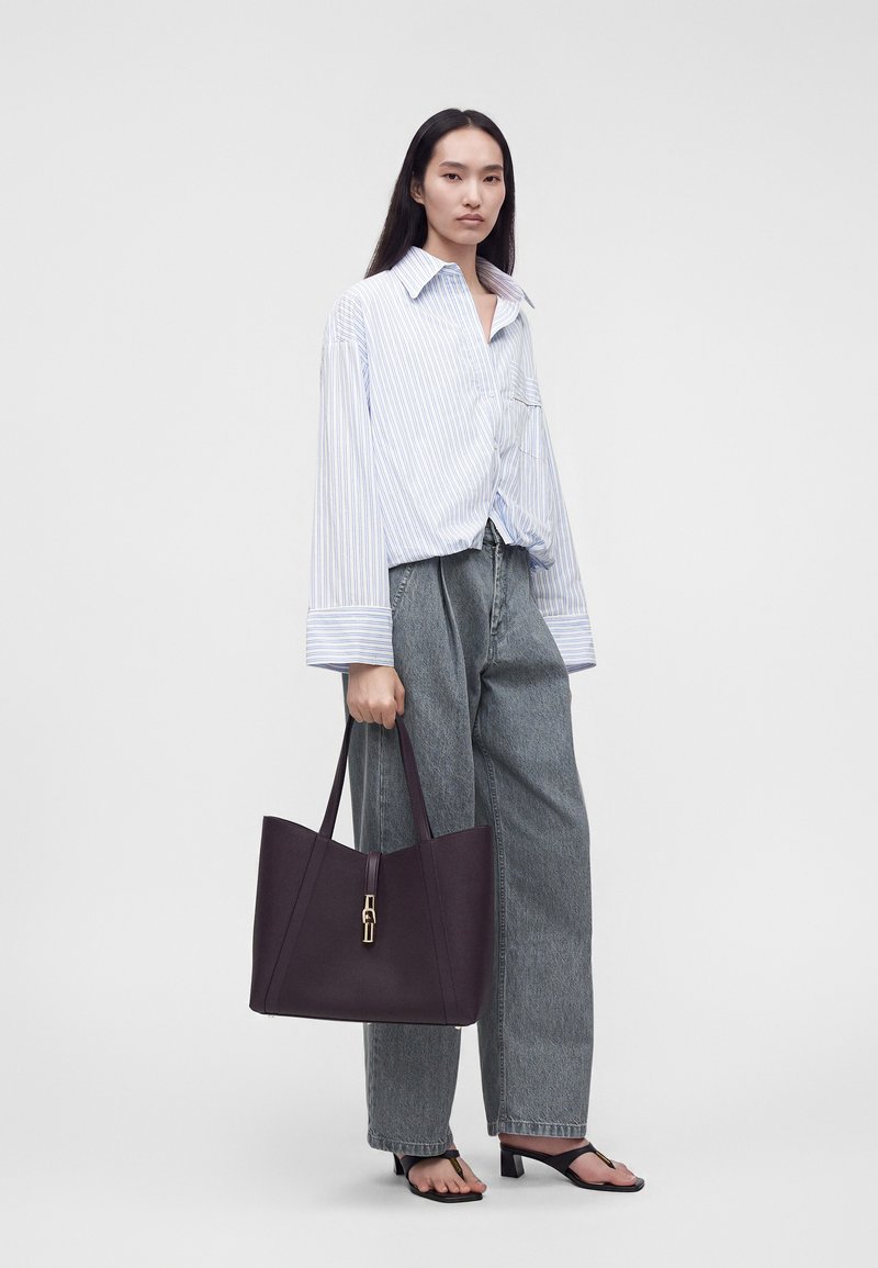 Femme portant une chemise blanche rayée oversize, un jean gris large, des sandales noires à talons, tenant un grand sac à main violet foncé, debout devant un fond uni.