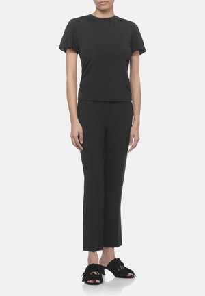 Donna che indossa una maglietta nera aderente a maniche corte, pantaloni neri a gamba dritta e sandali neri aperti con tacco e dettagli con frange.