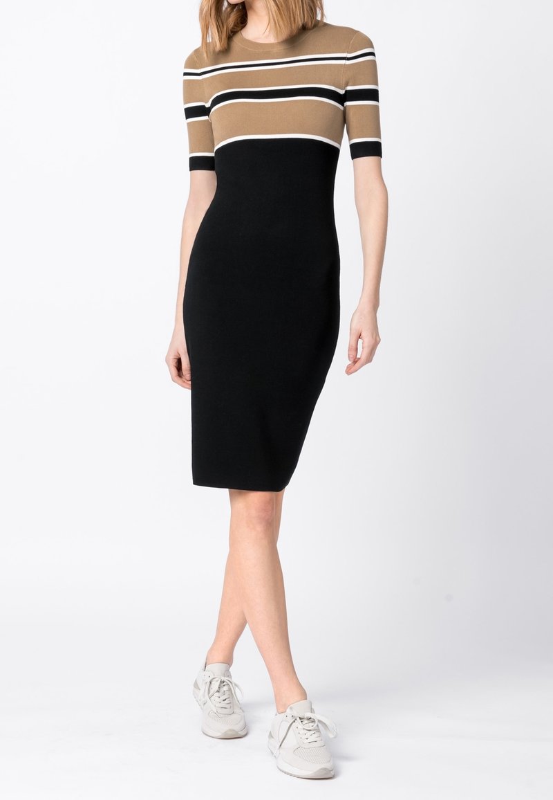 Femme portant une robe cintrée arrivant aux genoux avec des rayures horizontales beige, noires et blanches sur le haut, une jupe noire unie et des baskets blanches.