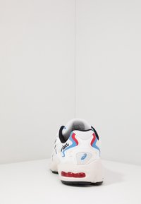 Vit sneaker med svarta accenter, blå och röda detaljer, mesh-överkörning, kraftig sula, vadderad krage och synlig hälkuddning.