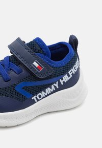 Modrá sportovní bota s vrchní vrstvou z meshových a syntetických materiálů, suchý zip a bílá gumová podrážka. Obsahuje bílé logo "TOMMY HILFIGER".