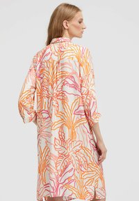 Frau mit langen Haaren, die ein lockeres, knielanges Kleid mit orange- und rosa-farbenen tropischen Blattmustern trägt, von hinten und von der Seite gezeigt.