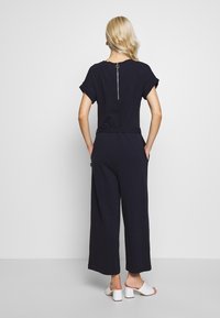 Marineblauer Jumpsuit mit kurzen, umgeschlagenen Ärmeln, lockerer Passform, Rückenreißverschluss und seitlichen Taschen. Kombiniert mit weißen Blockabsatzsandalen.