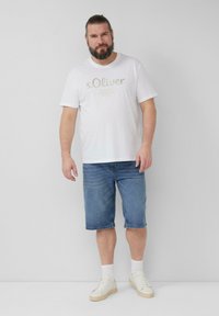 Biała bawełniana koszulka z jasnoróżowym logo, połączona z dżinsowymi szortami i białymi trampkami. Modelka stoi na neutralnym tle.