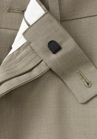 Detail opasku svetlohnedých nohavíc zobrazujúci gombík, gombíkovú dierku, zips a vnútornú bielu podšívku s hnedým pruhom.