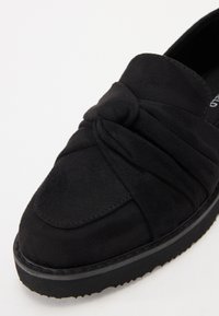 Svart nubuck slip-on sko med knuten frontdesign, rund tå, mjuk textur och tjock svart gummisula.