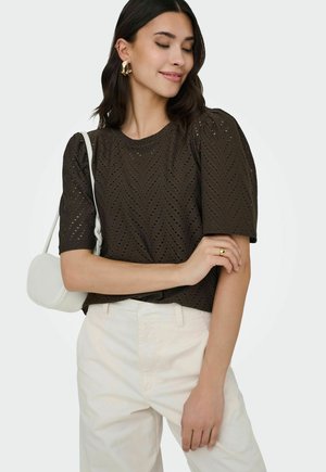 Vrouw draagt een donkerbruine eyelet top, crèmekleurige broek, gouden oorringen en ring, en draagt een witte schoudertas, poseert met gekruiste armen.