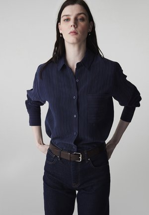 Kobieta o ciemnych włosach stoi z rękami na biodrach, ubrana w granatową koszulę na guziki z pionowymi paskami oraz ciemne dżinsy z brązowym paskiem.