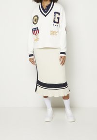 Sweat-shirt blanc avec des accents bleu marine et des patchs brodés, associé à une jupe crème côtelée avec un ourlet rayé bleu marine. Baskets blanches.