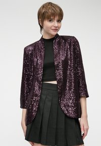 Jaqueta brilhante de lantejoulas roxa com mangas três quartos, frente aberta e textura suave, combinada com um top cropped preto e saia plissada.