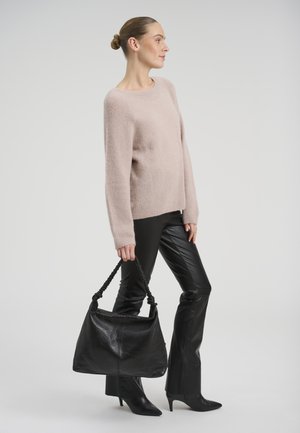 Nő bézs pulóverben, fekete bőrnadrágban és fekete hegyes sarkú cipőben áll, kezében nagy fekete táska fonott fogantyúkkal.