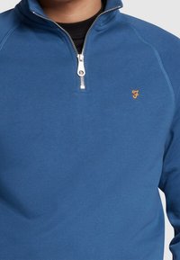Sweat-shirt à fermeture éclair bleu avec un col montant, des poignets côtelés et un petit logo orange sur la poitrine. Fabriqué en tissu doux et flexible.