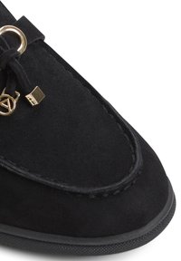 Scarpa in suede nera con forma arrotondata, caratterizzata da un accento dorato e un piccolo dettaglio in metallo. Finitura testurizzata e cuciture a contrasto.