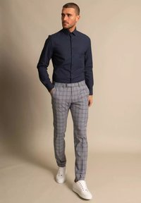 Mees, kellel on seljas tumesinine pikkade varrukatega nööbitav särk, hall ruuduline püksid ja valged tossud, seisab ühe käega taskus.