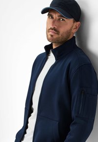 Navy zip-up jacket made of soft fabric, featuring a high collar, side pockets, and left arm pocket. Worn with a matching cap.