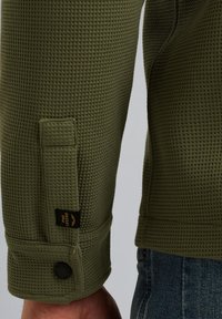 Olijfgroen gestructureerd sweatshirt met een wafelbreisel, voorzien van een knoopmanchet en een merklabel op de mouwzoom.