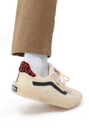 Beżowe trampki Vans z czarnym paskiem i czerwonym tyłem w panterkę, noszone z białą skarpetką i brązowymi spodniami na białym tle.