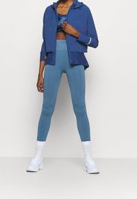 Blå idrottsjacka med huva över en marinblå camouflagesportbehå. Matchande blå leggings och vita sneakers med vita ankelsockor.