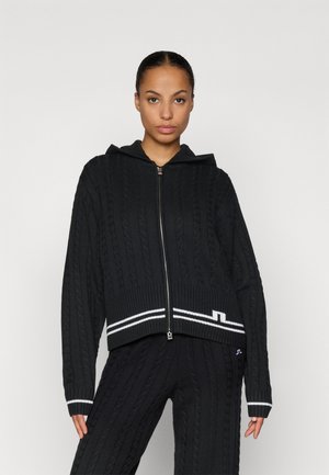 Black cable-knit hooded sweater with a zip closure, featuring white stripe accents at the hem and cuffs. Soft texture and relaxed fit.