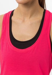Jasně růžový sportovní top s žebrovanou texturou, vrstvený přes černý top. Kolo výstřih, lehký materiál vhodný na cvičení.