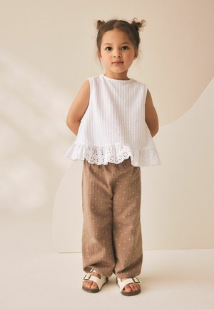 Young girl with hair in buns wearing white sleeveless top with ruffle hem, brown polka dot pants, and white sandals, standing with hands behind back.