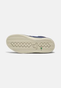 La semelle de la sneaker présente un caoutchouc beige texturé avec un motif ondulé, un accent de logo vert et une pointe arrondie. Un design durable et fonctionnel.