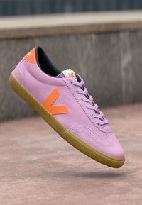 Ružové semišové tenisky s oranžovým gumovým akcentom a logom. Vyznačujú sa nízkym profilom, texturovanými šnúrkami a gumovou podrážkou.