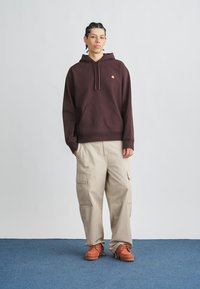 Felpa con cappuccio marrone con tasca frontale e logo giallo, abbinata a pantaloni cargo beige con tasche laterali e scarpe arancioni. Sfondo semplice.