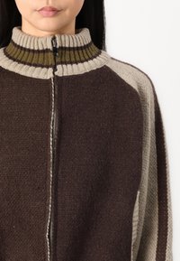 Brown and beige knit zip-up sweater with a high collar, featuring contrasting textured stripes and a silver zipper detail.